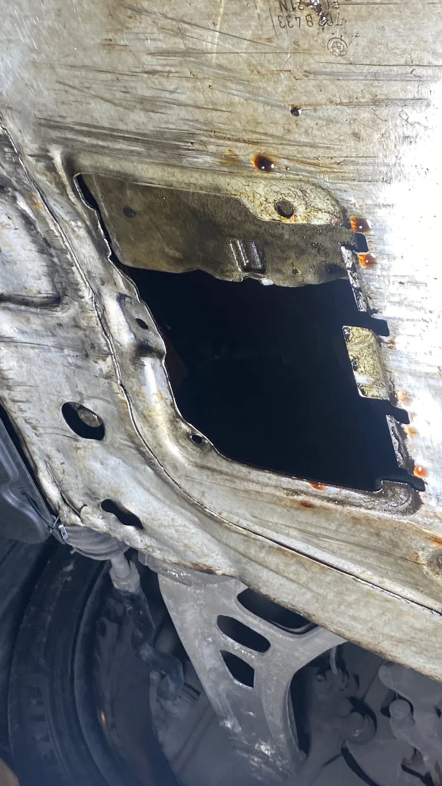 Rectangular hole rusted through the underbody floor panel with orange rust drips, from below