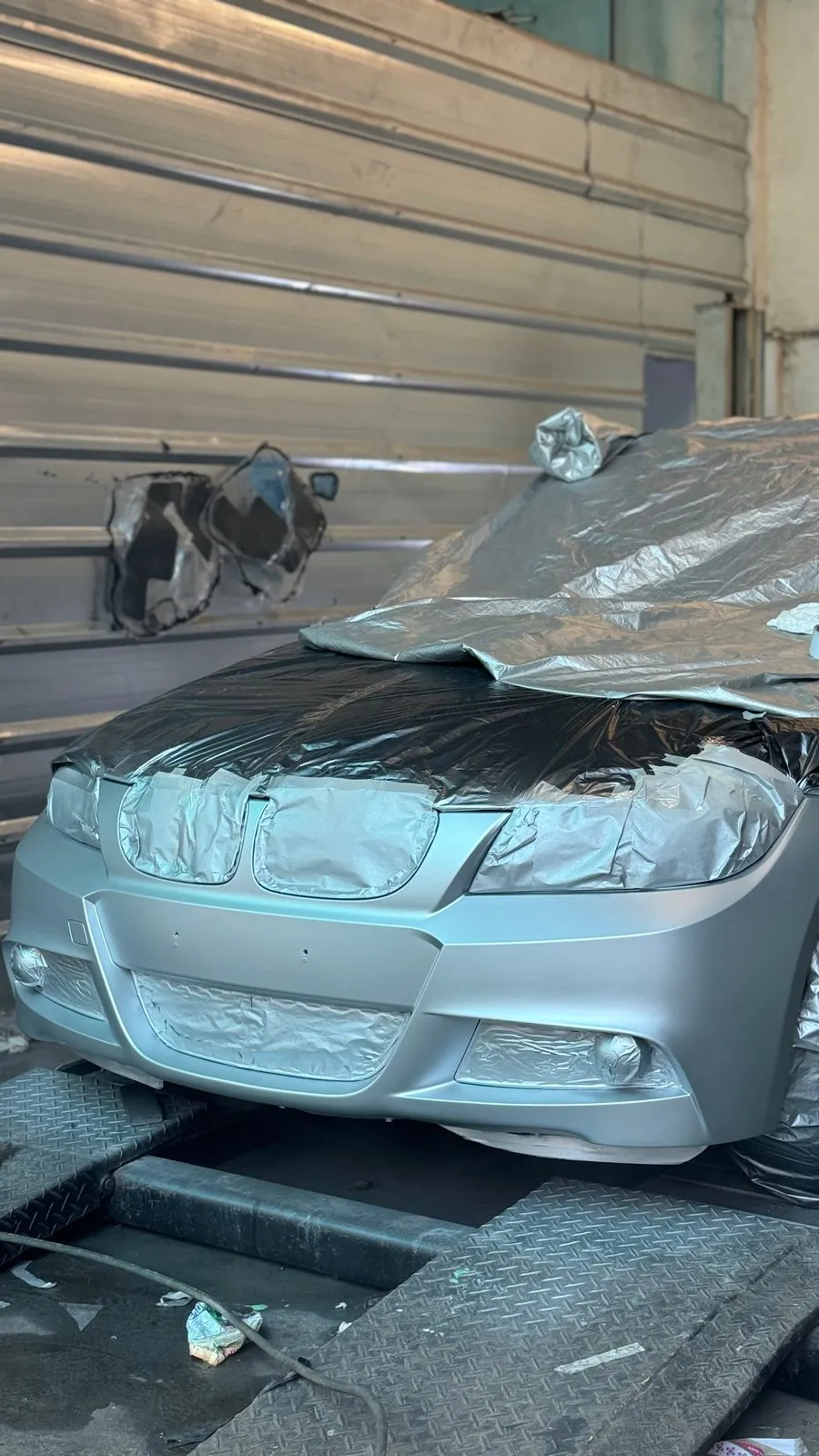 Front of the silver E90 covered in grey plastic masking with the bonnet and bumper in matt primer, in the workshop